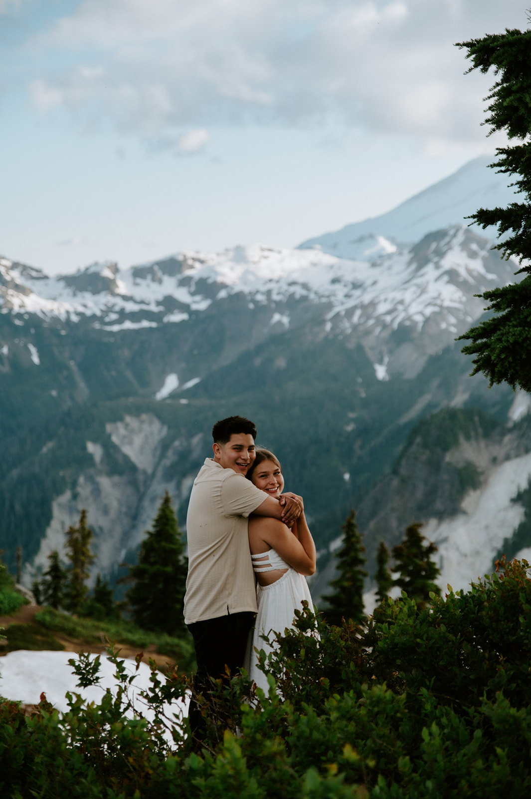 Couple hugging and smiling together on a mountain overlook surrounded by evergreen trees and alpine views