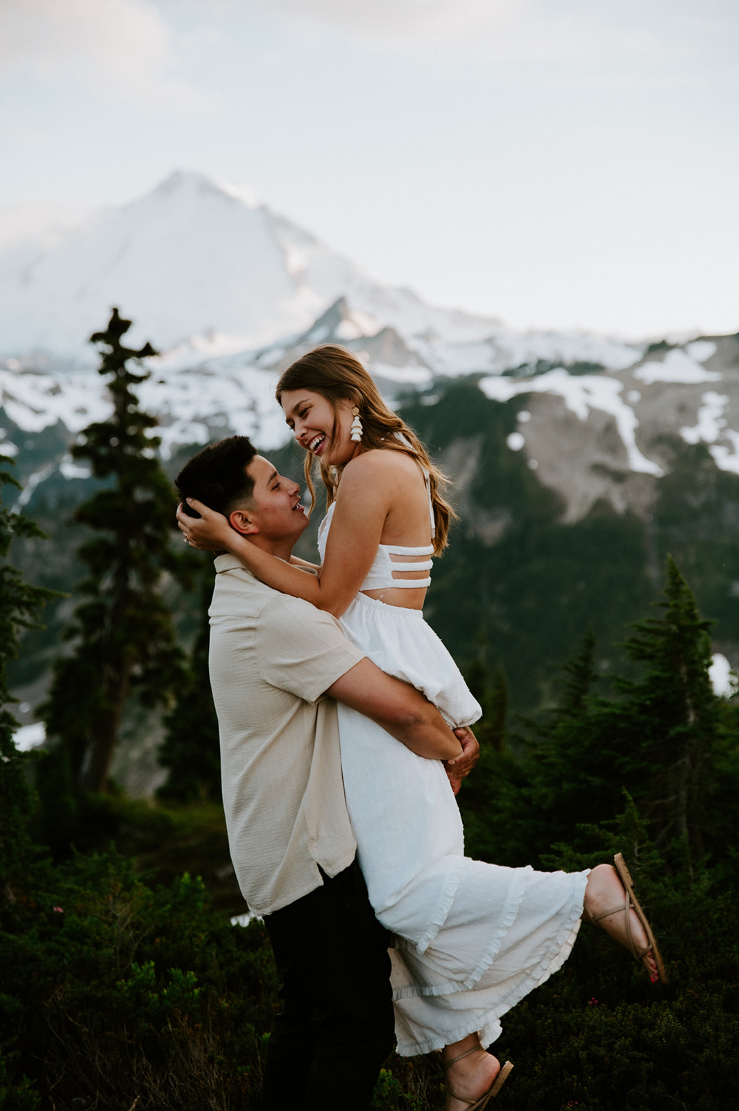 Romantic mountain engagement photoshoot with groom lifting bride-to-be in front of alpine views and evergreen trees
