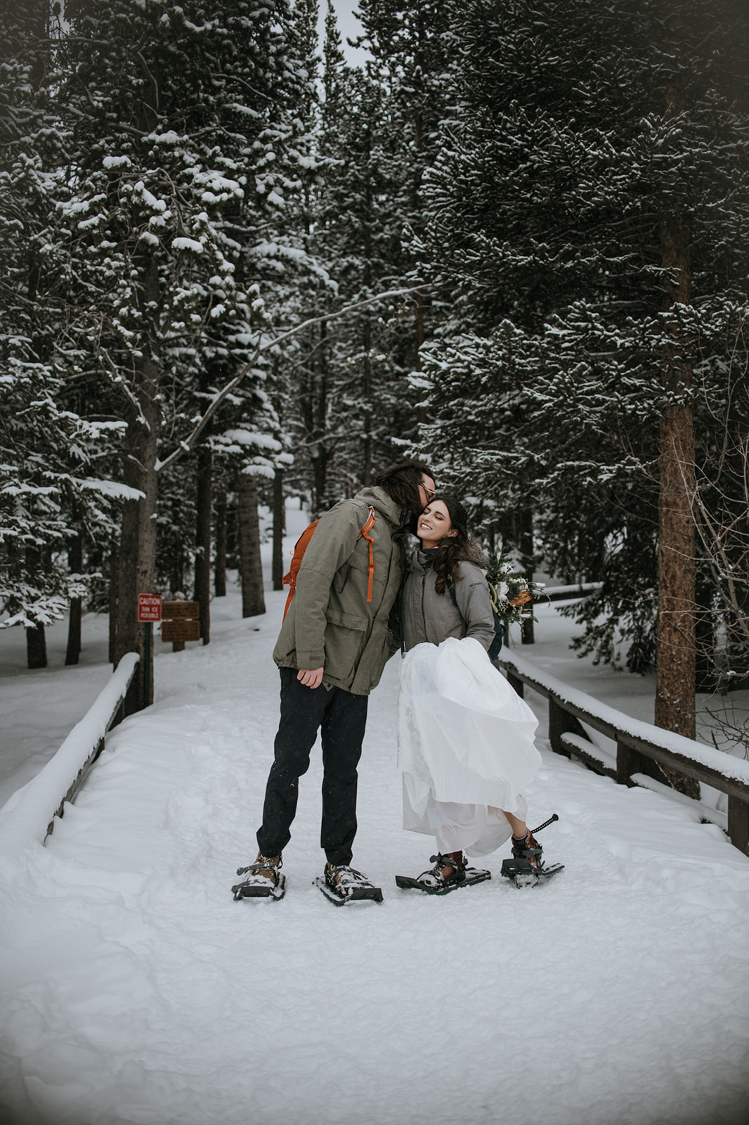 Playful winter engagement photoshoot with couple laughing together on a snowy trail wearing snowshoes