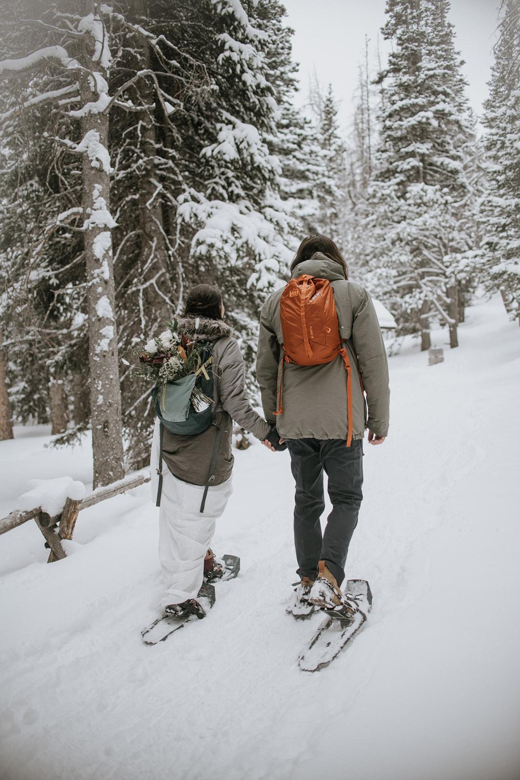 Couple walking hand in hand through a snow-covered forest trail wearing backpacks and snowshoes