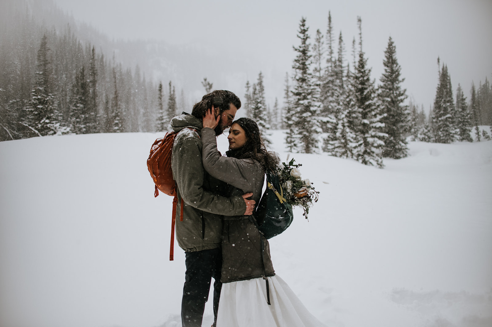 Cozy winter engagement photoshoot with couple embracing in falling snow with pine trees in the background