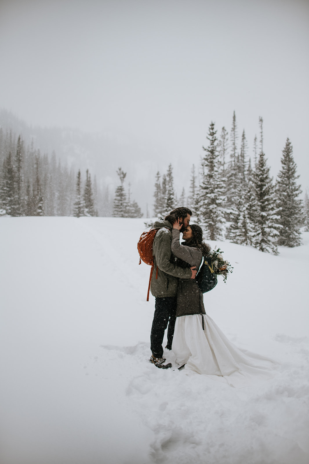 Romantic winter engagement photoshoot with couple kissing in a snowy landscape with pine trees and falling snow