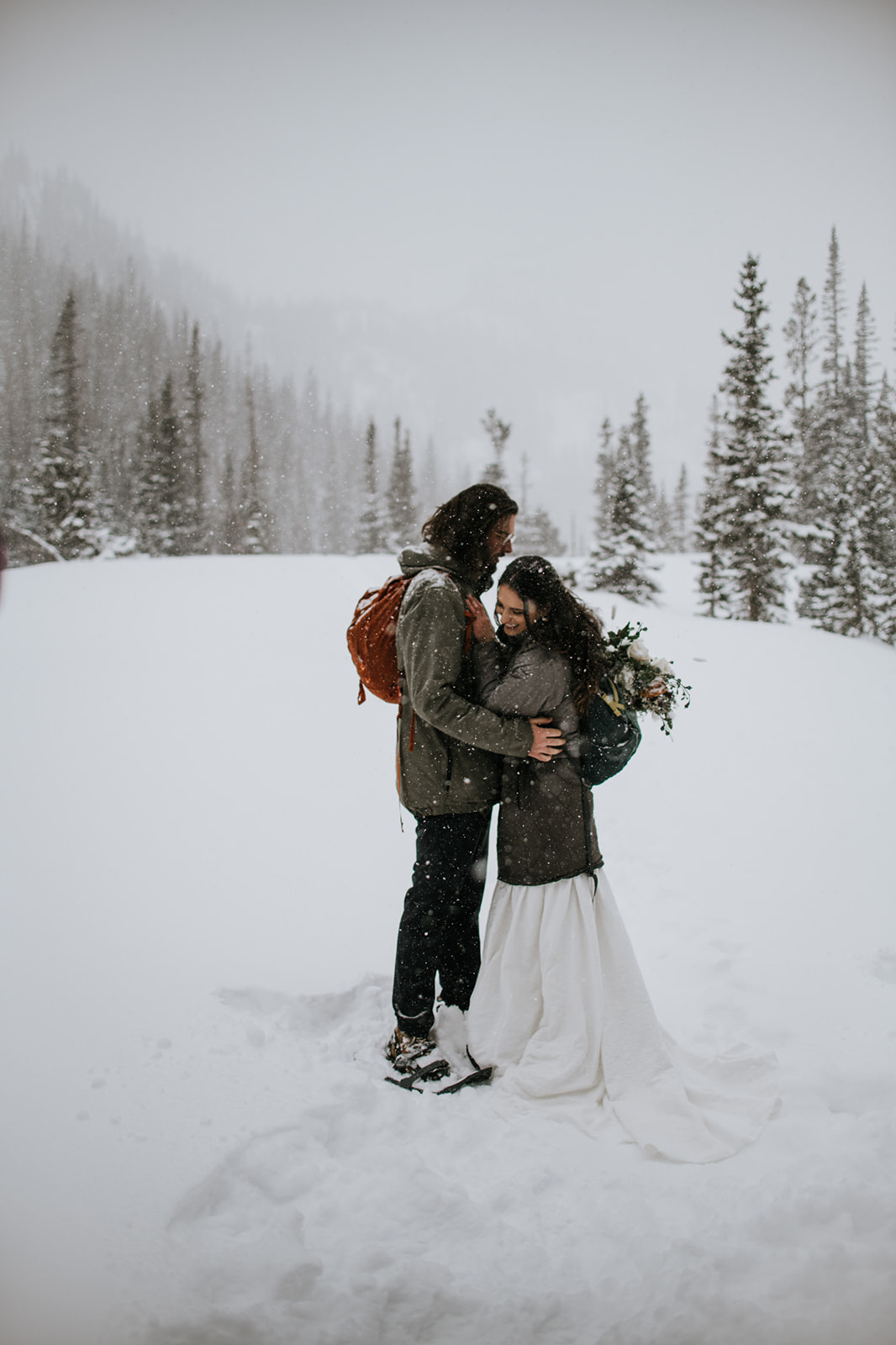 Cozy winter engagement photoshoot with couple embracing in falling snow surrounded by pine trees and soft fog
