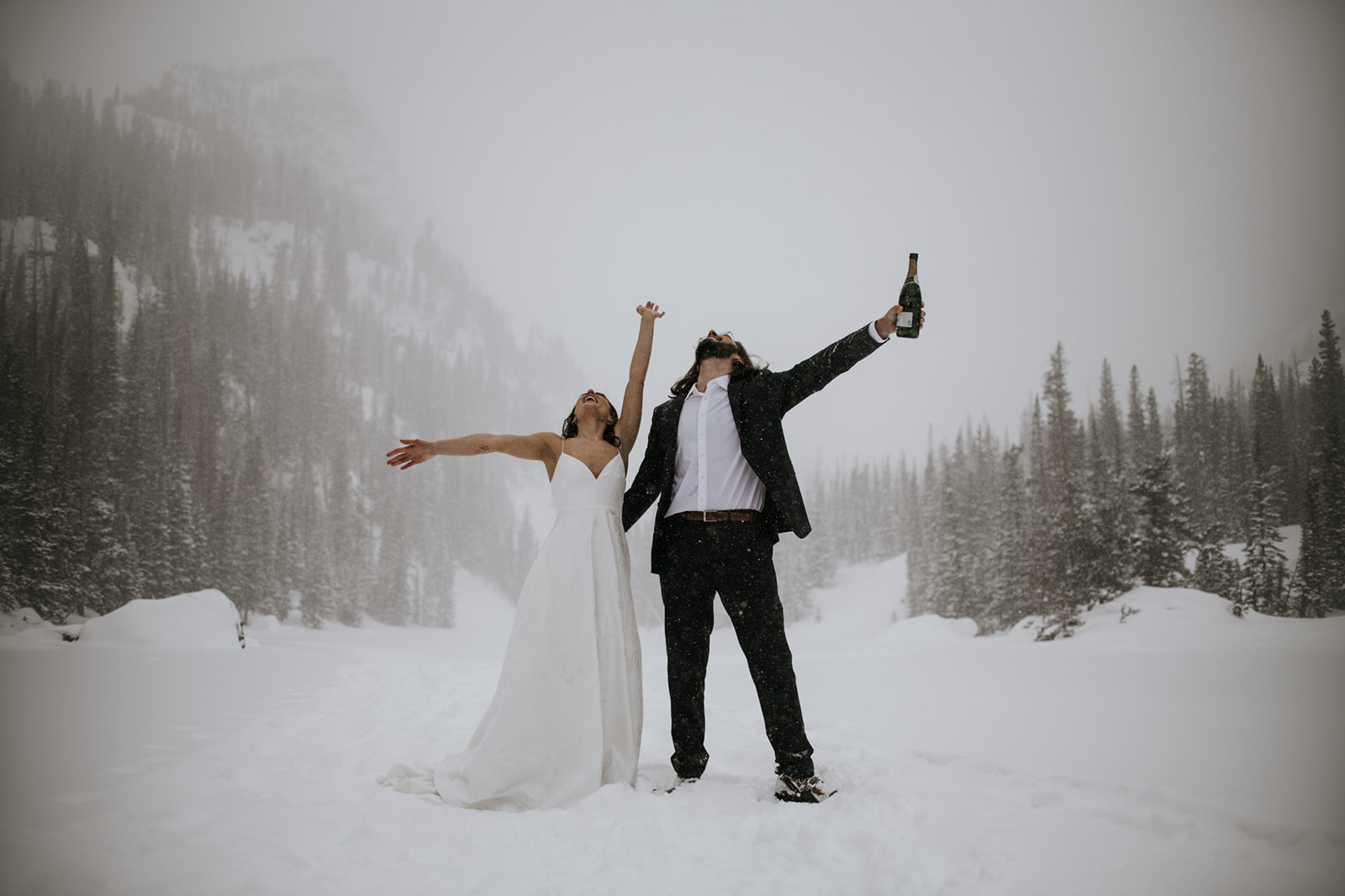 Joyful winter engagement photoshoot in falling snow with couple celebrating in an open mountain landscape