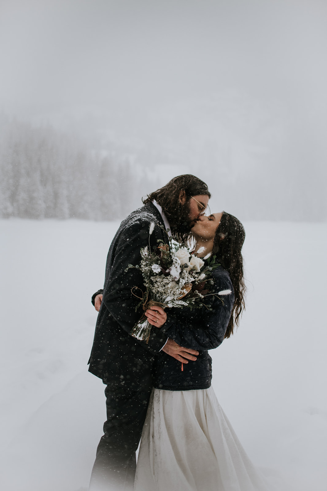 Intimate snowy engagement photoshoot with couple kissing while holding a bouquet in a winter landscape