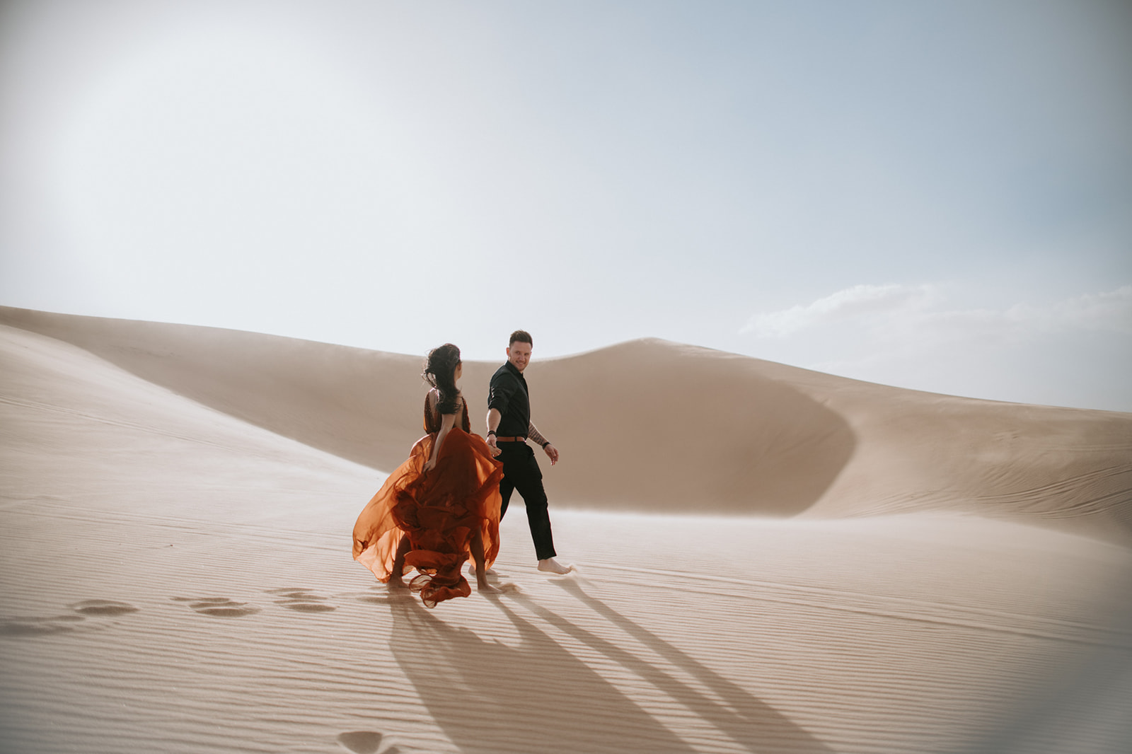 Couple walking hand in hand across desert dunes with long shadows stretching behind them