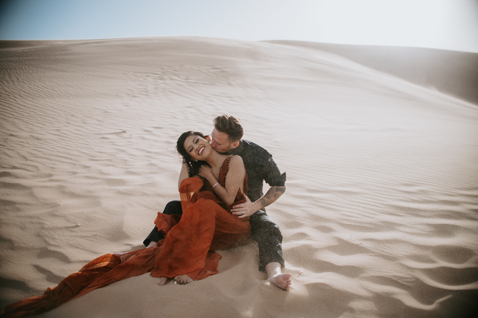 Joyful engagement photoshoot with couple laughing together while sitting in textured sand dunes