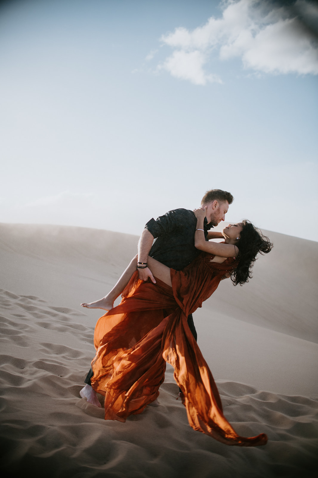 Couple embracing and dipping in flowing dress on sunlit sand dunes at golden hour