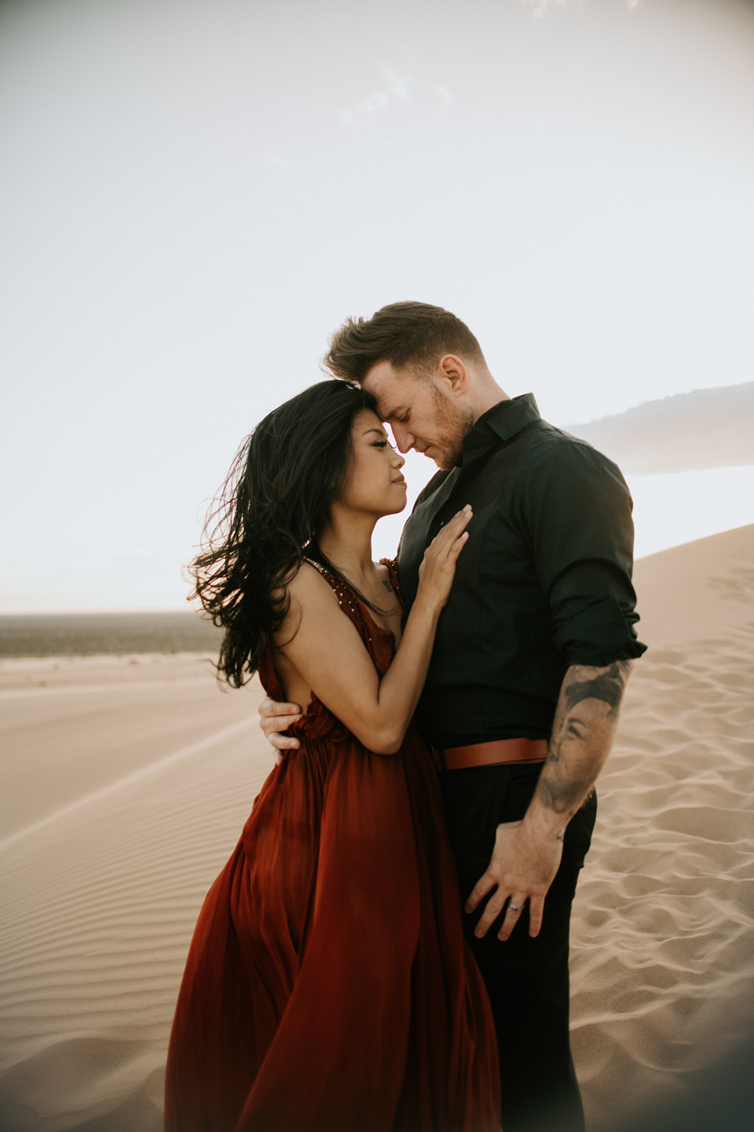 Close-up of couple embracing on sand dunes with soft golden light and wind-swept dress
