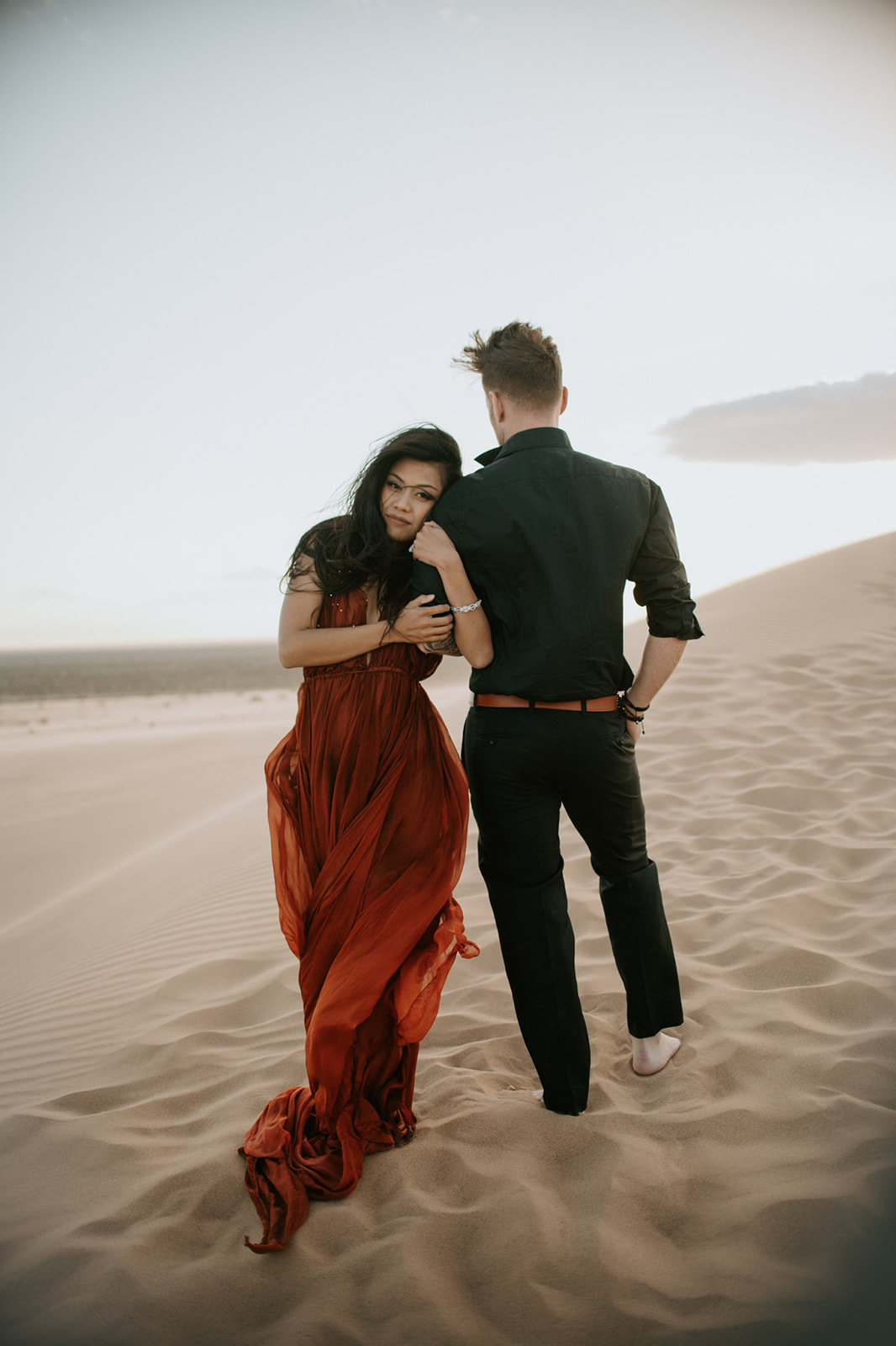 Intimate desert engagement photoshoot with couple walking through sand dunes at sunset, dress trailing behind