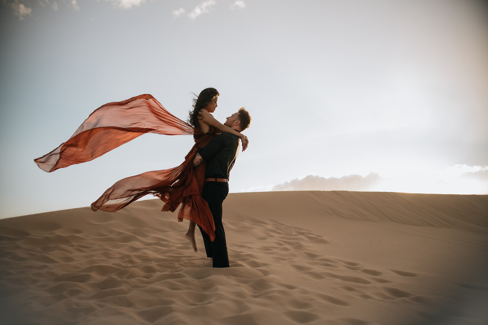 Romantic engagement photoshoot in the desert with partner lifting bride-to-be as her dress blows in the wind