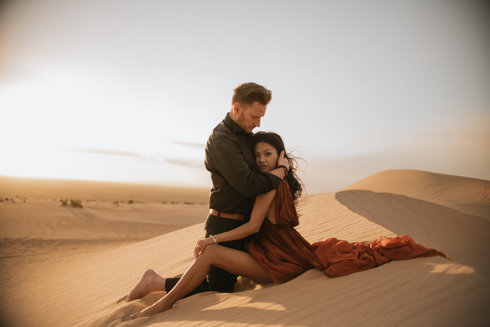 Romantic desert engagement photoshoot with couple cuddling on sand dunes at sunset