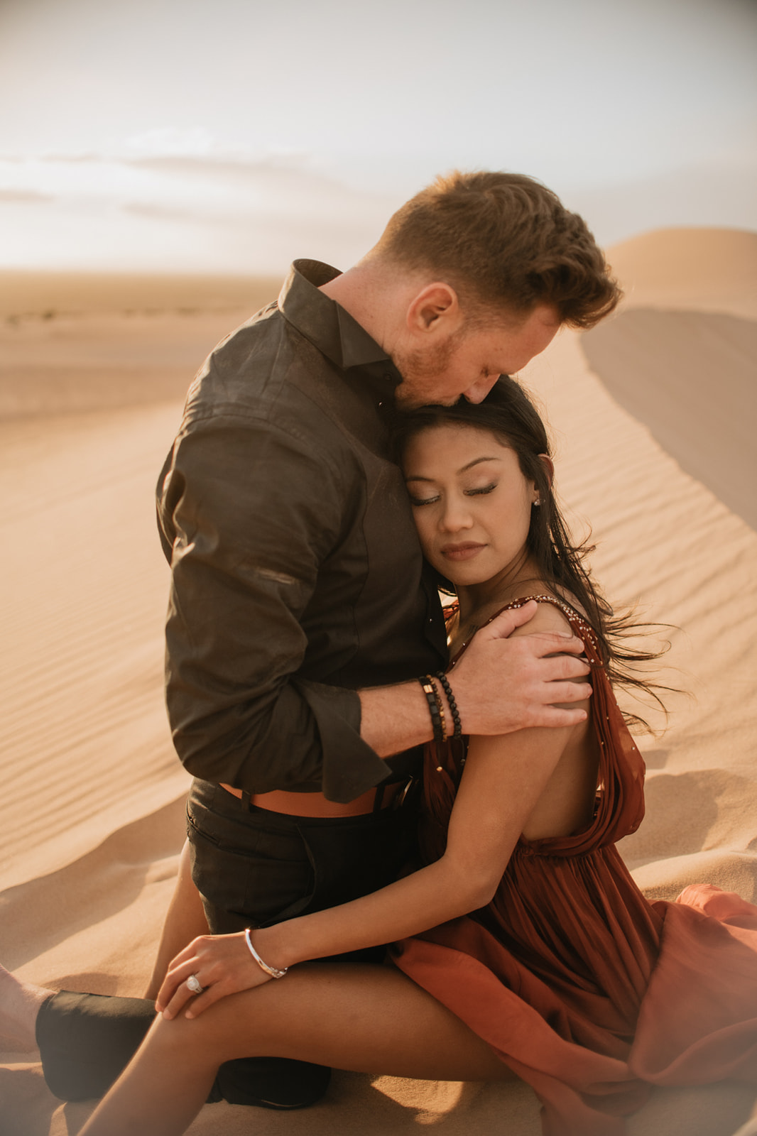 Intimate moment with couple cuddling on sunlit dunes, warm tones and soft shadows creating a romantic feel