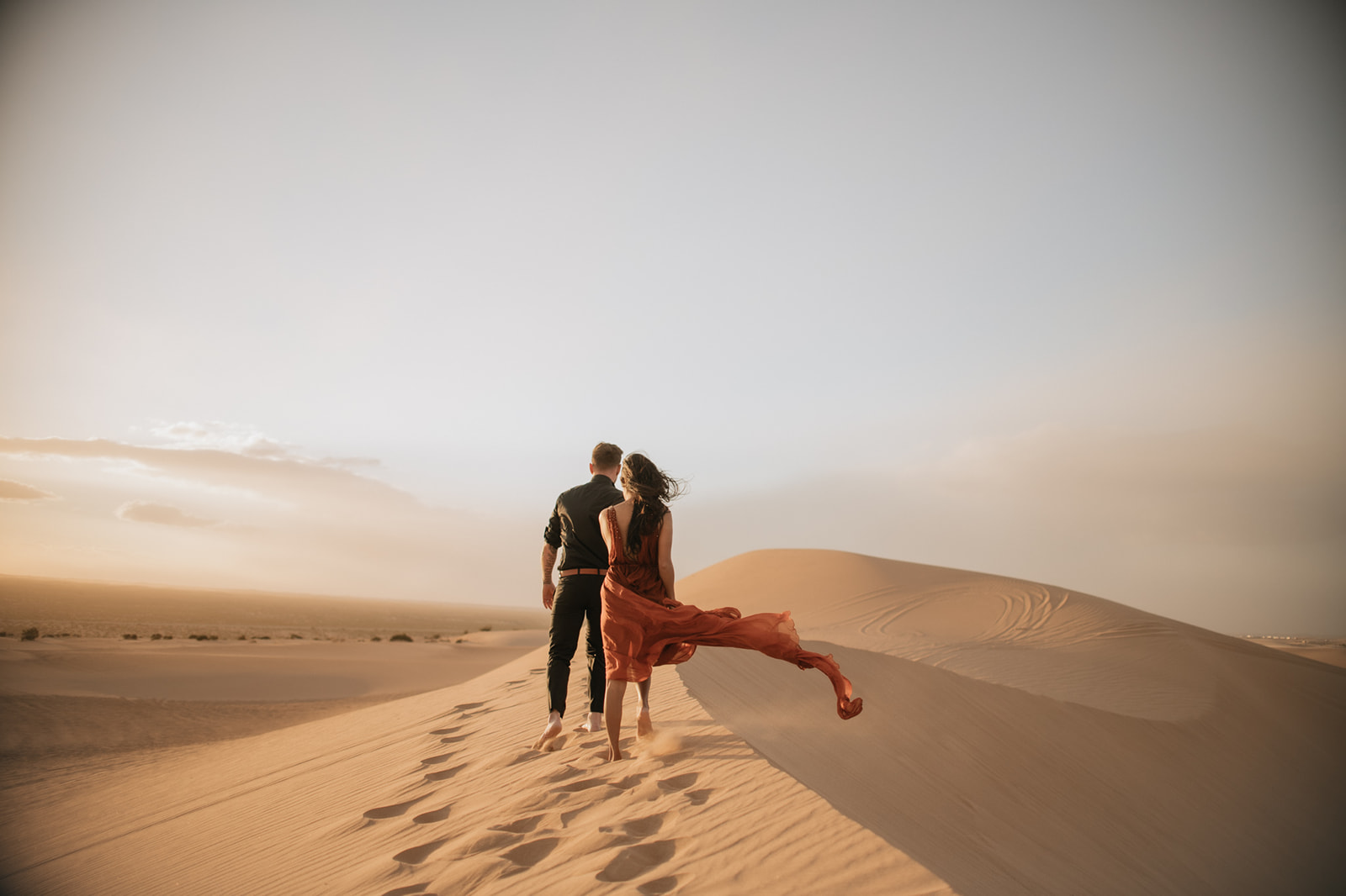 Couple walking barefoot along golden sand dunes with flowing dress trailing behind at sunset
