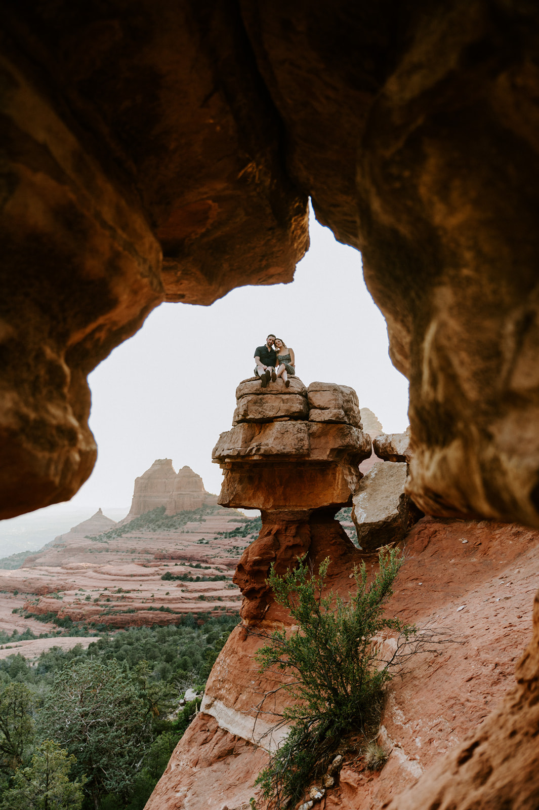 Wide shot of a couple sitting on a rock formation framed by a natural arch after they elope to Sedona