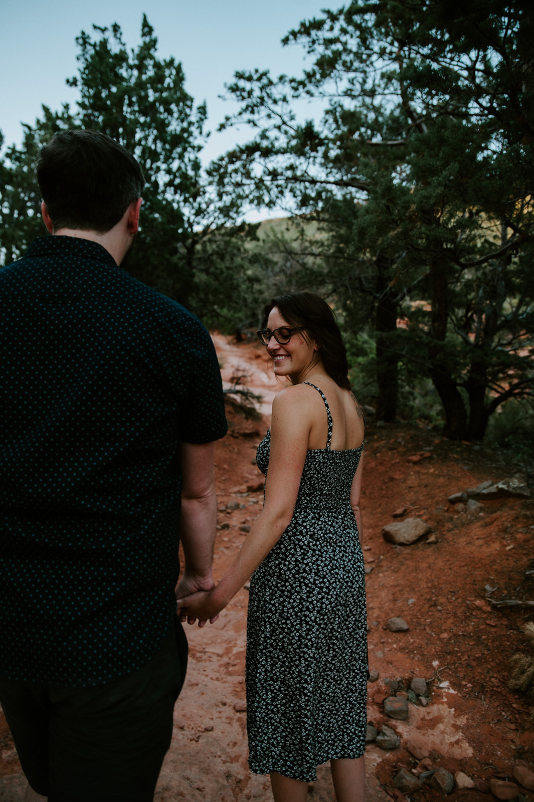 Couple walking hand in hand along a red dirt trail surrounded by trees in Sedona