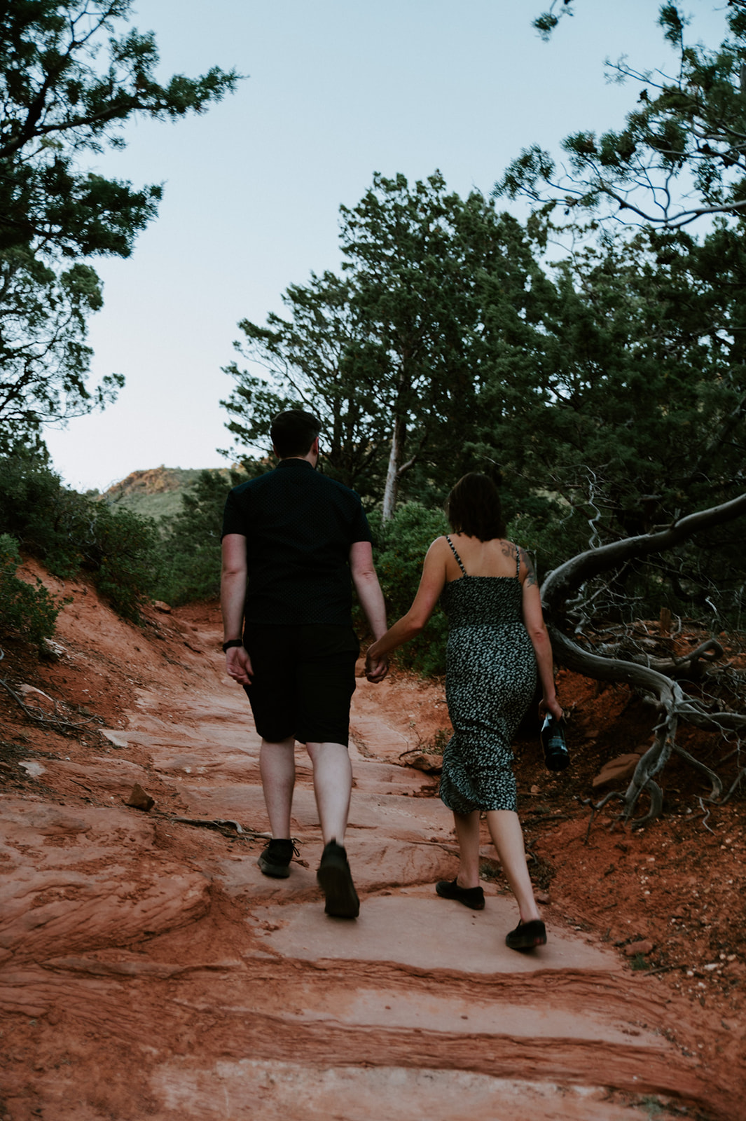 Couple walking hand in hand up a red rock trail surrounded by trees in Sedona