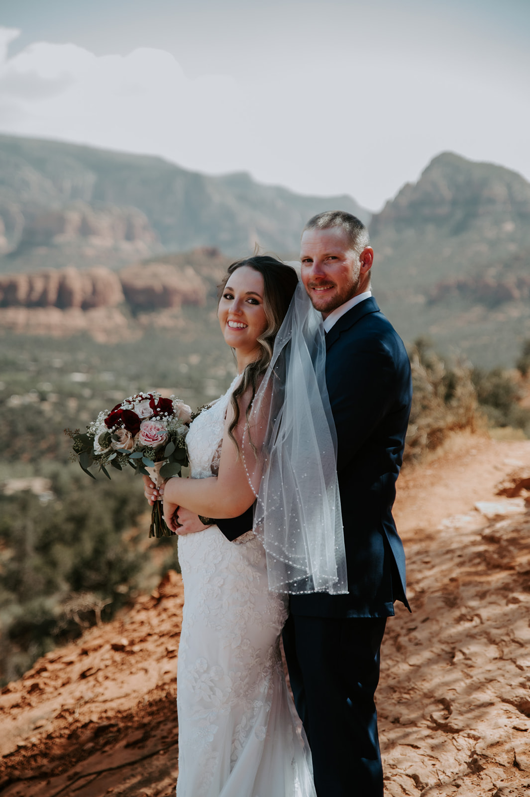 Bride and groom standing together on a red rock overlook with mountains in the distance after they elope to Sedona
