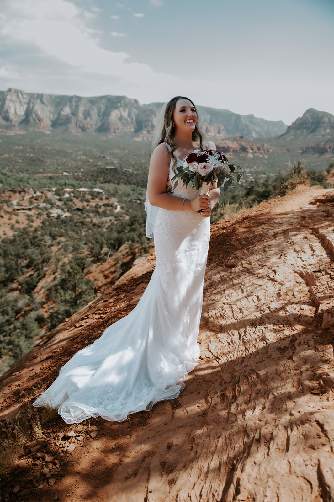 Bride standing on a red rock slope holding her bouquet with Sedona mountains in the background after she elopes to Sedona