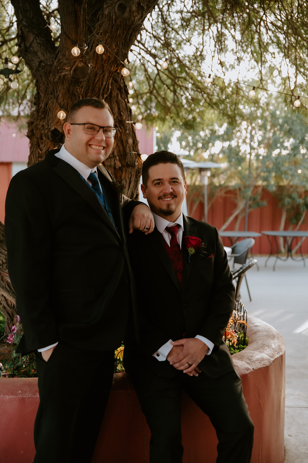 Two grooms smiling together beneath string lights before their outdoor wedding ceremony