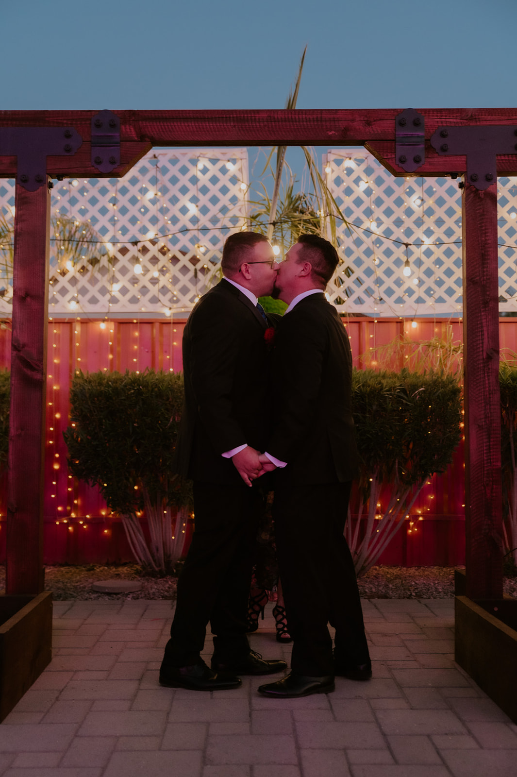 Same-sex couple kissing beneath a wooden ceremony arch during a romantic evening wedding photographed by an LGBTQ wedding photographer