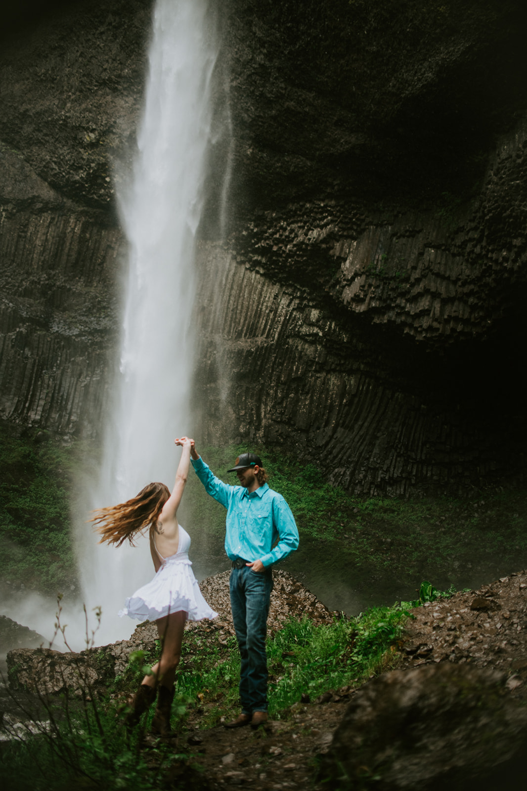 Couple spinning beneath a waterfall surrounded by lush greenery and rugged Oregon cliffs.