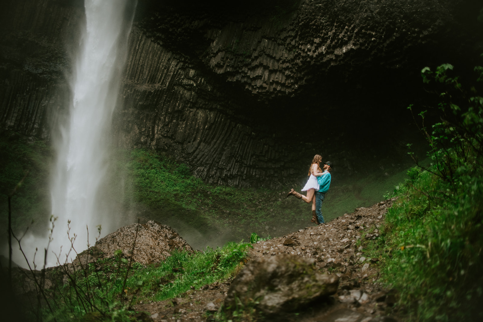 Romantic waterfall portrait in Oregon with couple embracing beneath dramatic basalt cliffs and mist.
