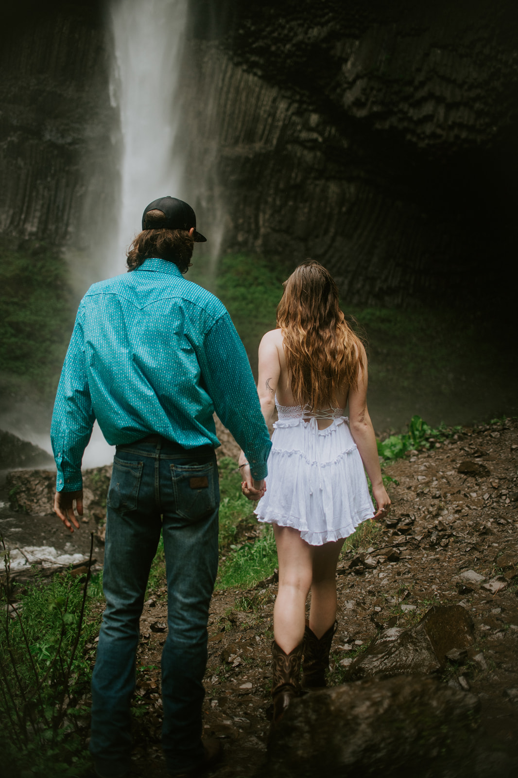 Couple walking hand in hand toward a waterfall during a rainy forest session in Oregon.