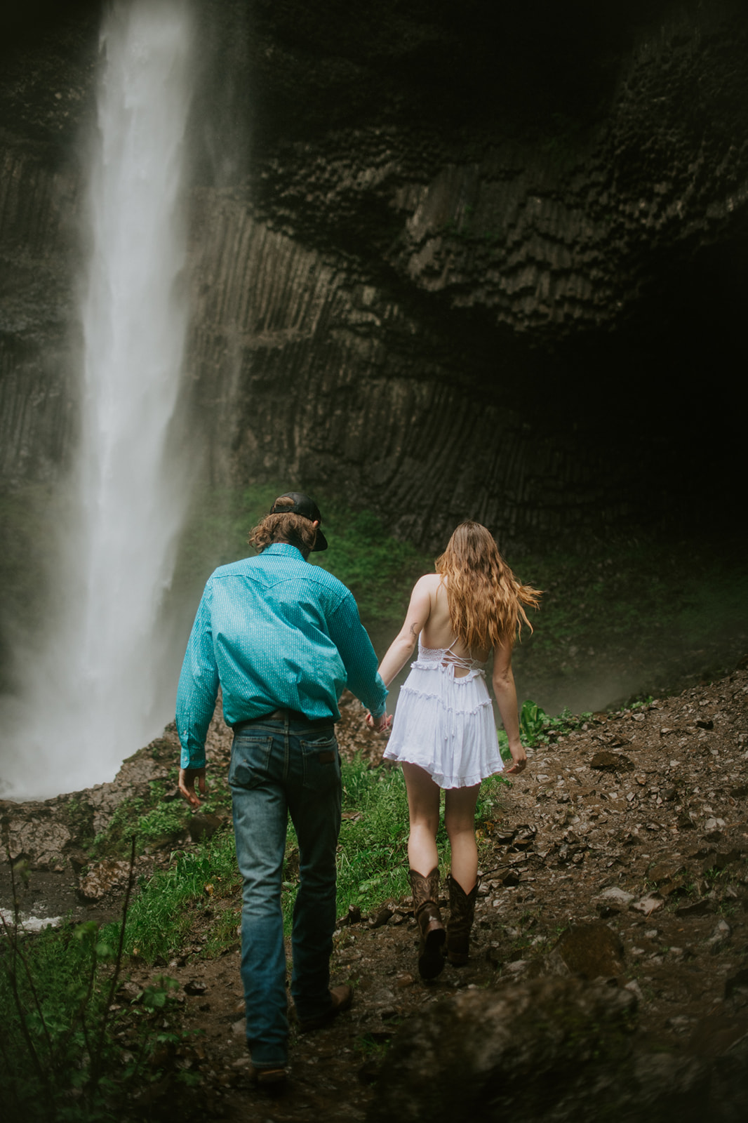Adventurous couple hiking toward a dramatic waterfall during a rainy forest session in Oregon.