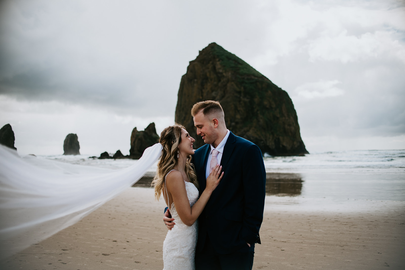 Bride and groom smiling together on the Oregon Coast with flowing veil and moody beach scenery.