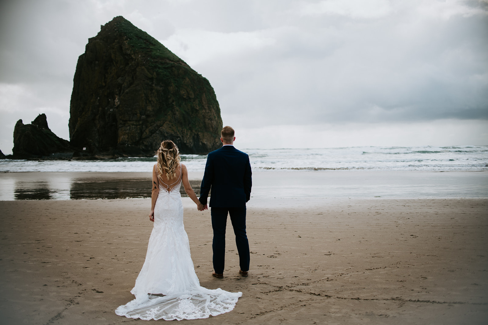 Bride and groom standing hand in hand at Cannon Beach with Haystack Rock and stormy skies behind them.