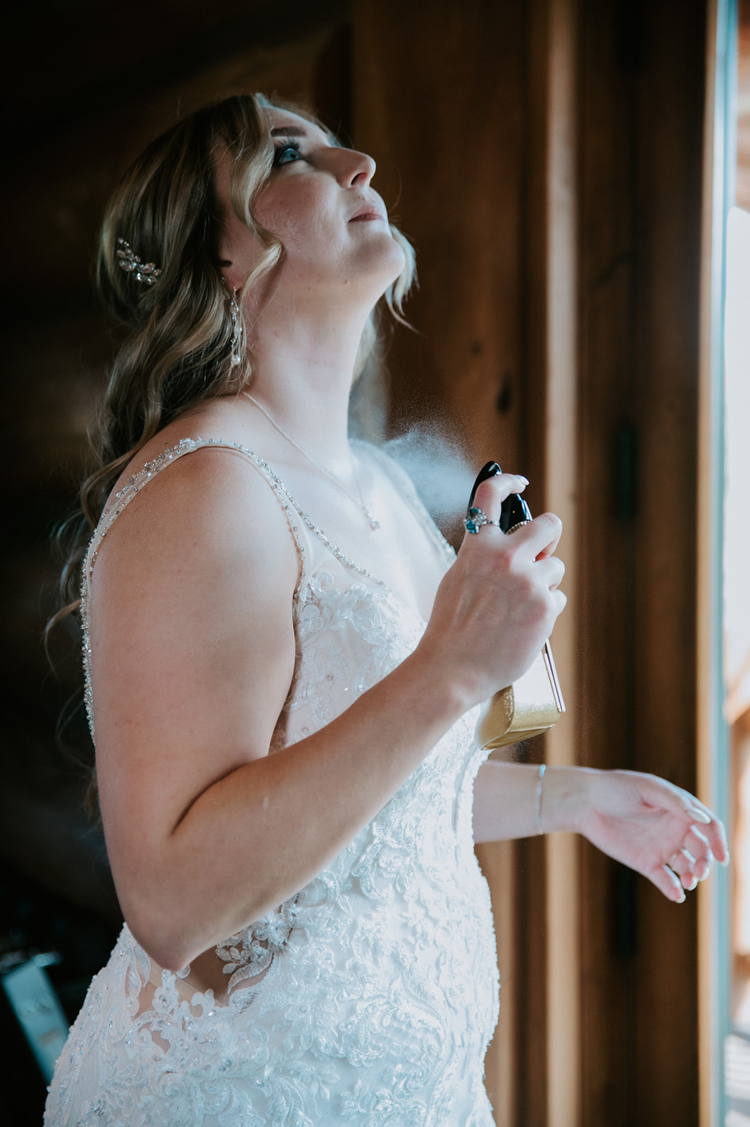 Bride sprays perfume near a window while finishing her wedding day preparations.
