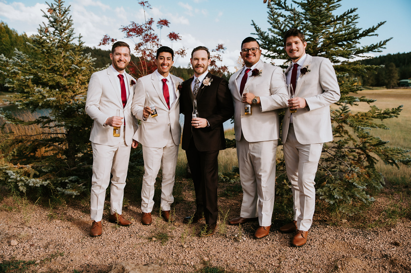 Groom and groomsmen pose together outdoors with drinks in hand before the wedding reception, celebrating with friends in a mountain setting.
