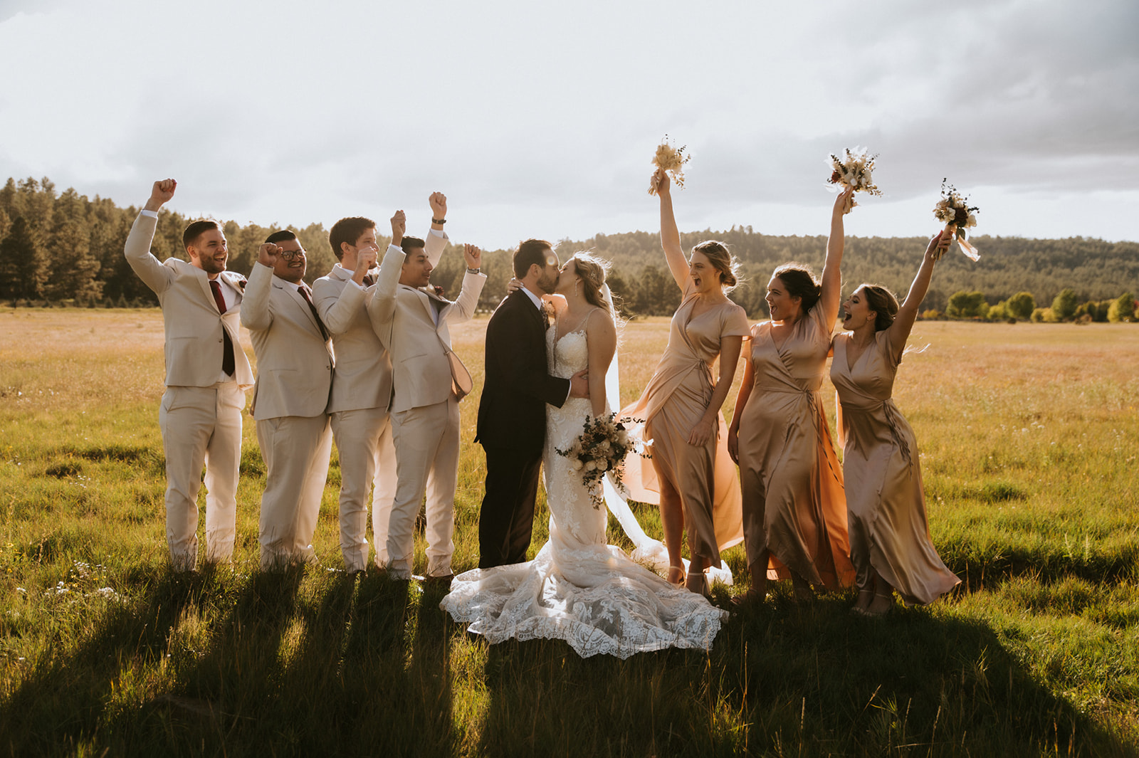 Wedding party celebrates as the newlyweds share a kiss in an open meadow at sunset.
