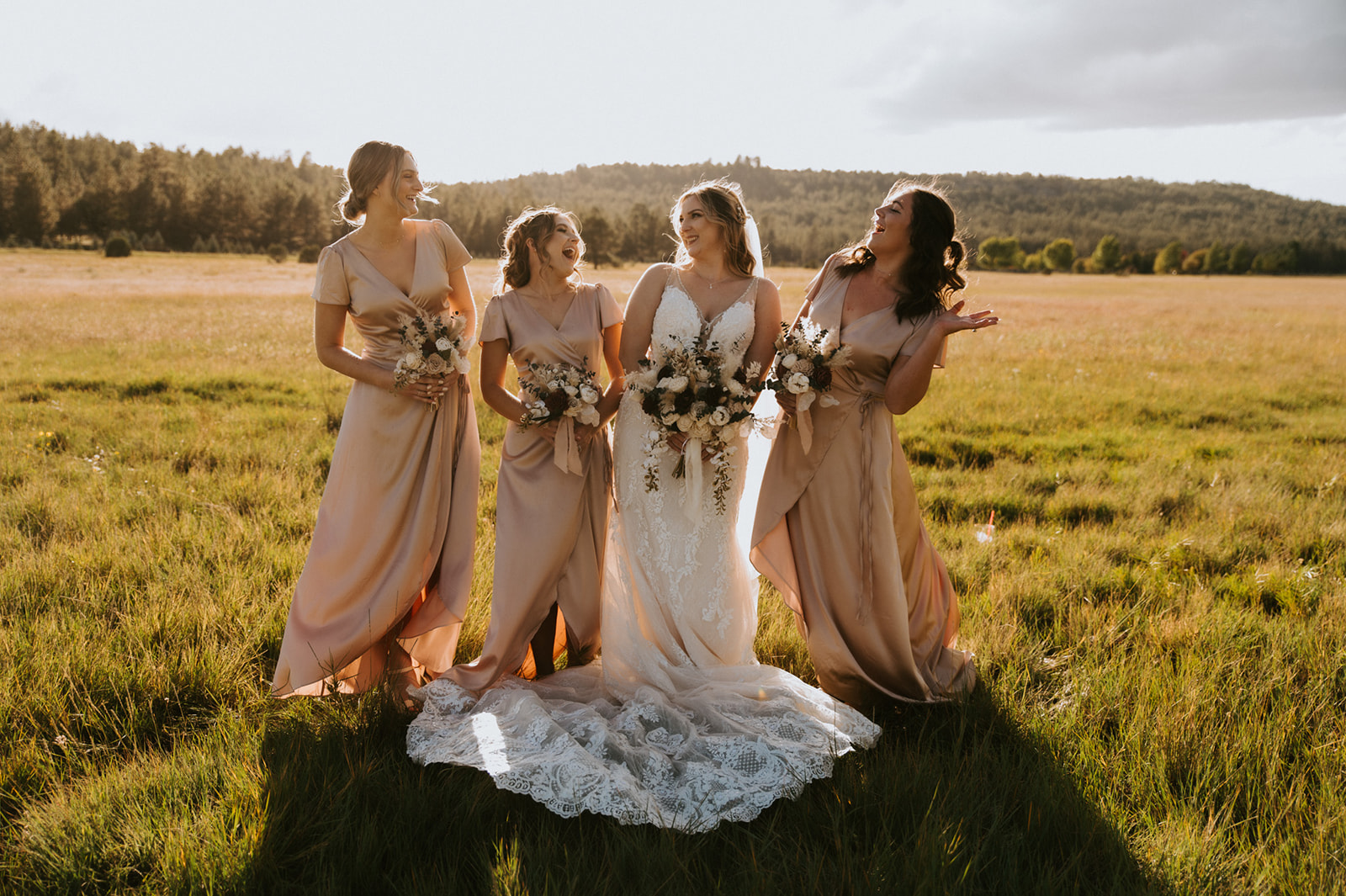 Bride stands with her bridesmaids in a sunlit meadow, holding bouquets and sharing laughter on the wedding day.
