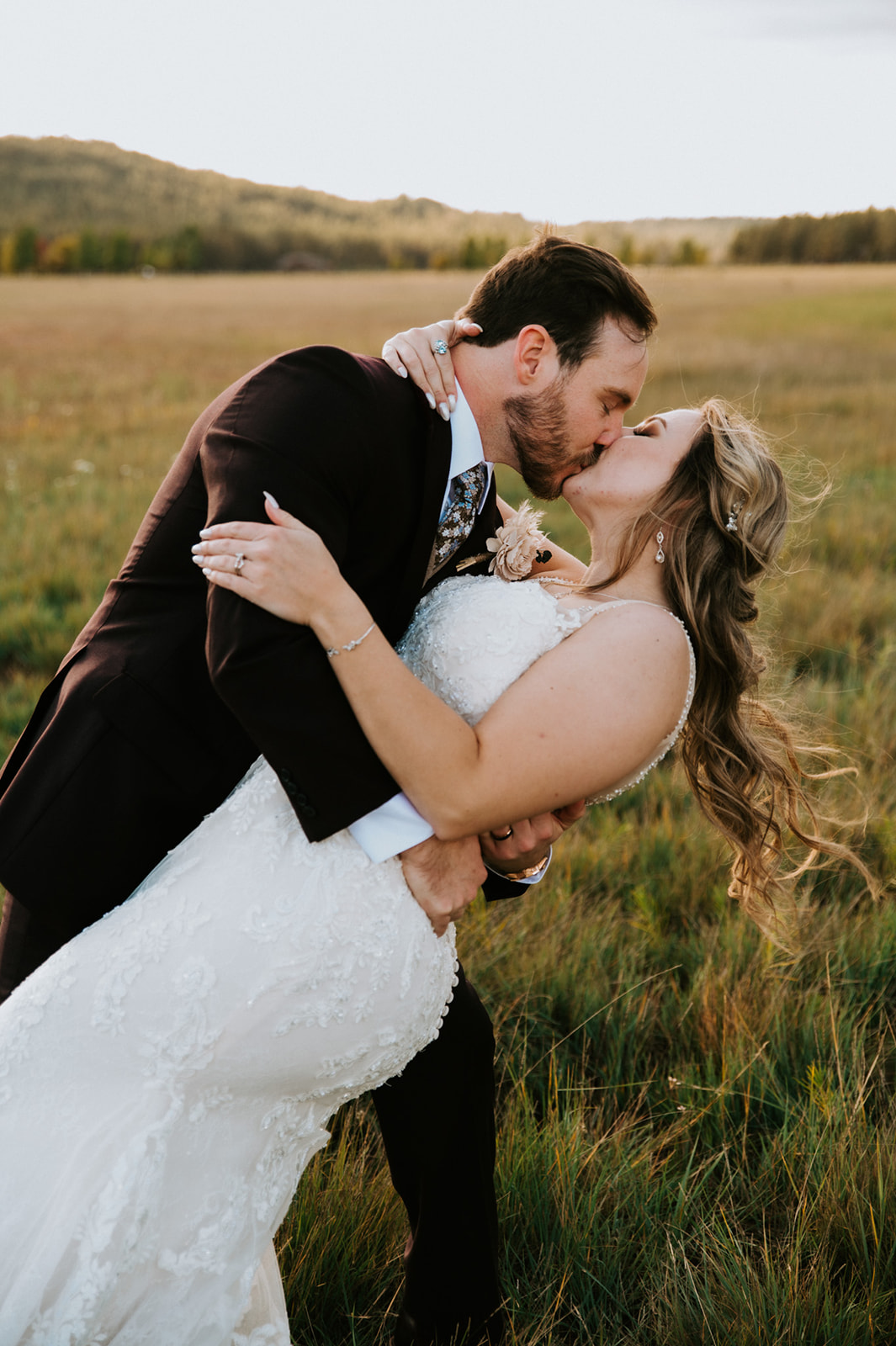Bride and groom share a dramatic dip kiss in an open field at golden hour, surrounded by mountain views.
