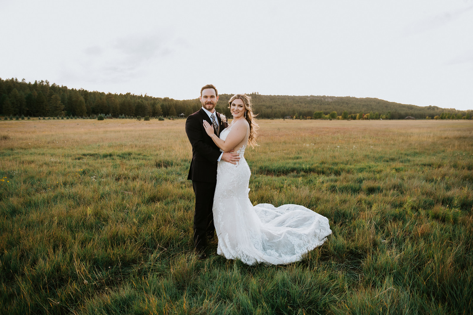 Newly married couple pose together in an open meadow at sunset, showcasing a relaxed and natural wedding portrait.
