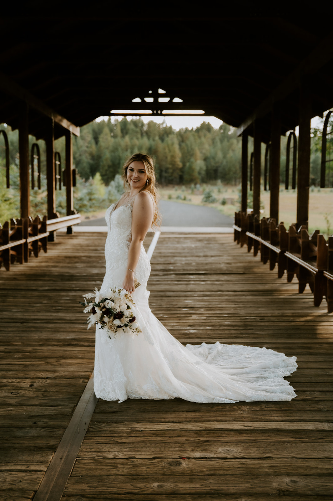 Bride poses alone on a covered wooden bridge with her bouquet and lace train displayed behind her.
