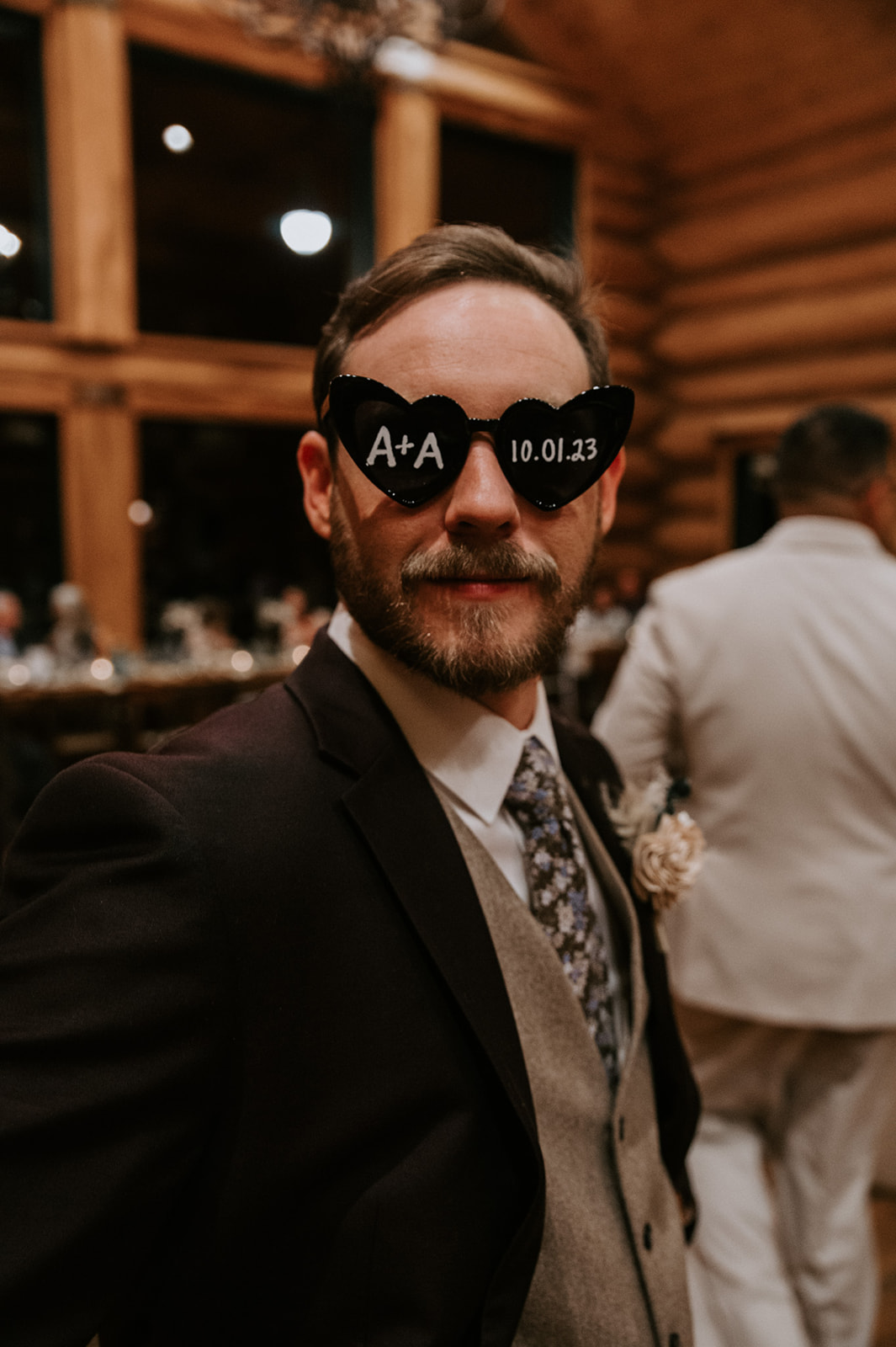 Groom wearing custom wedding sunglasses poses during the reception inside a rustic timber-frame venue.
