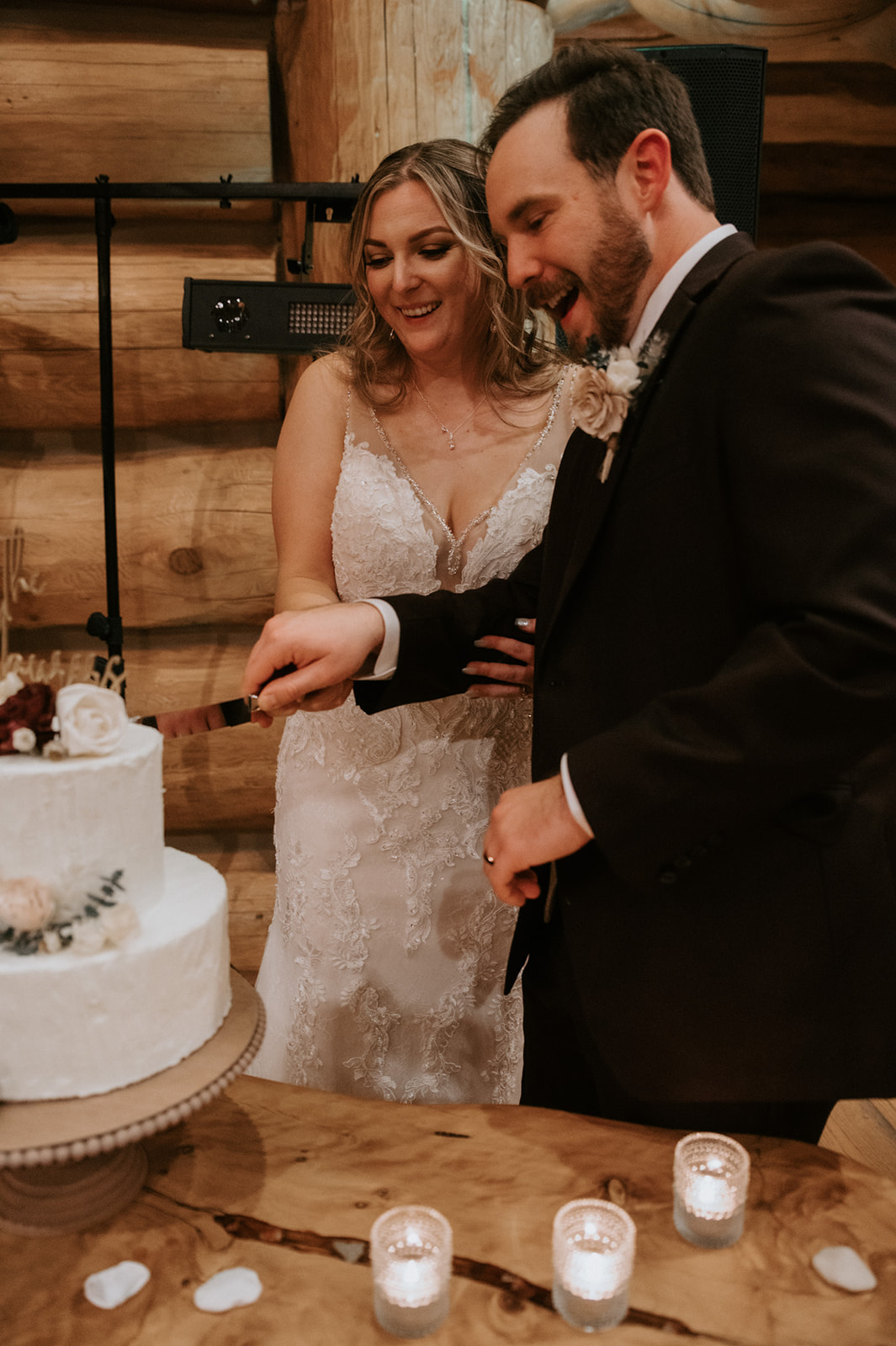 Bride and groom smile together while cutting their wedding cake at a cozy wood-paneled reception venue.
