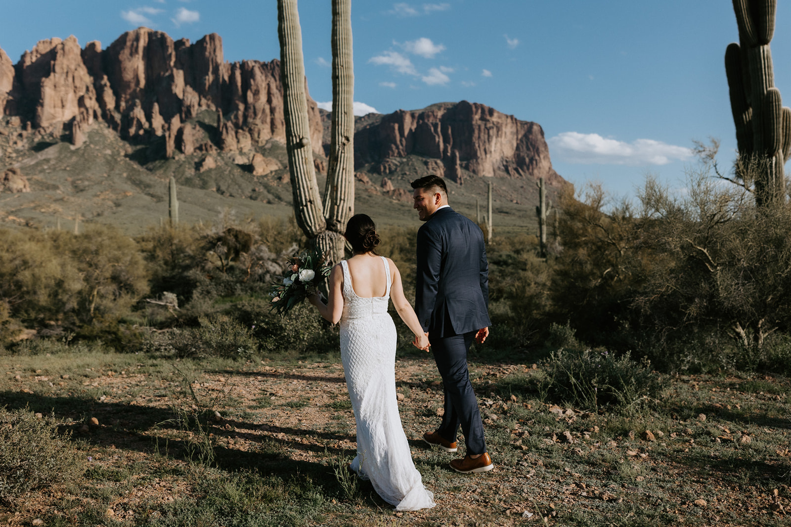 Bride and groom walking hand in hand through the Arizona desert surrounded by saguaros and mountain views after eloping in Arizona.