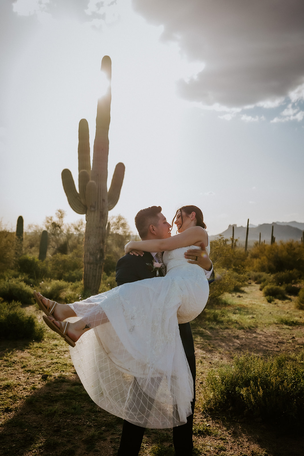 Groom carrying bride beneath a towering saguaro cactus during golden hour while eloping in Arizona desert landscape.