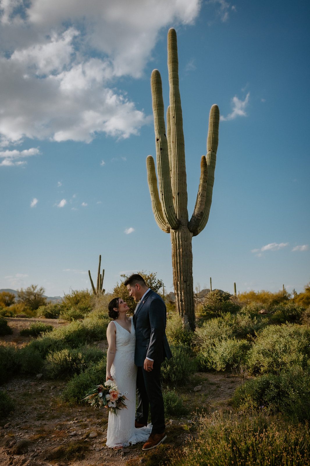 Romantic portrait of a couple standing beneath a giant saguaro cactus in the Arizona desert at golden hour
