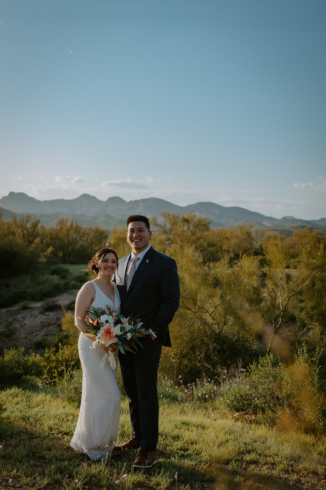 Bride and groom smiling together with desert mountains and wildflowers surrounding their Arizona wedding portraits
