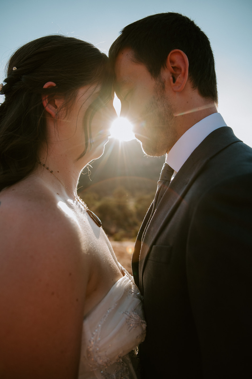 Close-up portrait of a bride and groom touching foreheads with sunlight glowing between them
