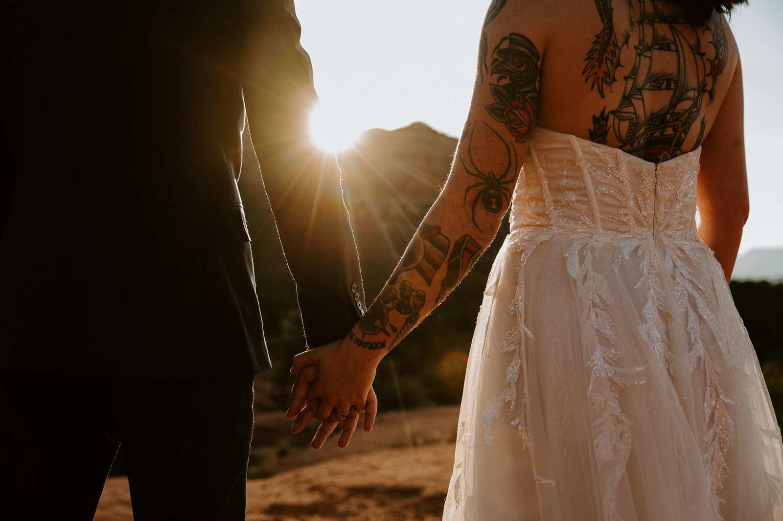 Tattooed bride and groom holding hands at golden hour with sun flare shining across the desert landscape
