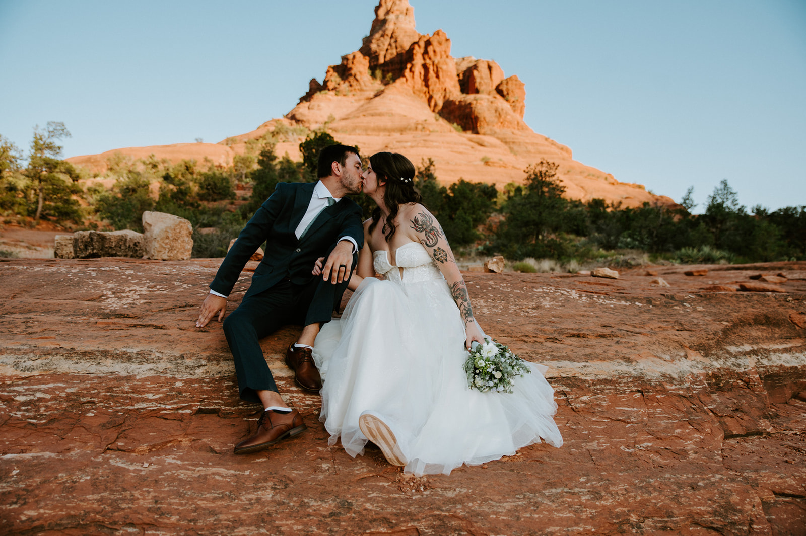 Couple sharing a kiss on red rock terrain after eloping in Arizona with Bell Rock visible in the background.