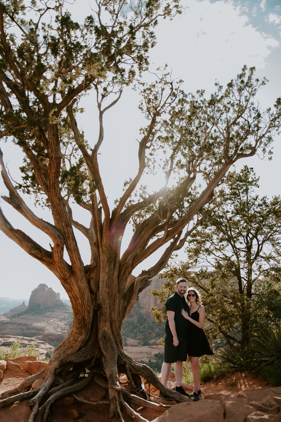 Couple standing beneath a large juniper tree overlooking Sedona's iconic red rock landscape during an engagement session.