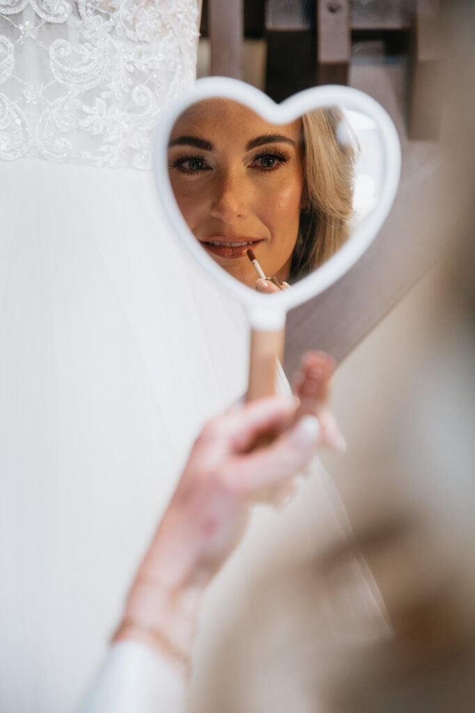 Bride applying lipstick while looking into a heart-shaped mirror during the getting-ready portion of her wedding day.