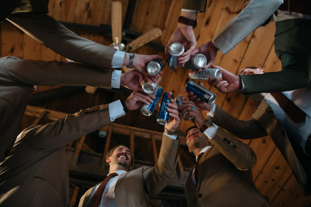 Groomsmen raising beer cans for a celebratory toast while getting ready inside a mountain cabin before the wedding.